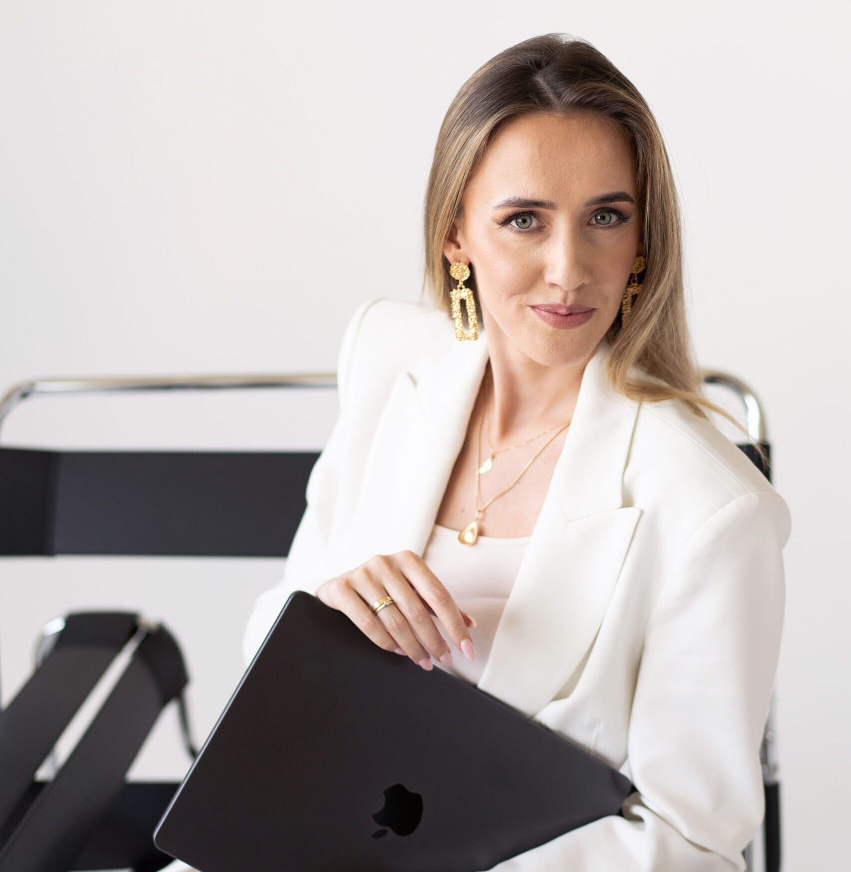 Dagmara Grzeszczuk z The Sparkling Media od marketingu dla architektów siedzi na czarnym metalowym krześle i ma na sobie biały garnitur.