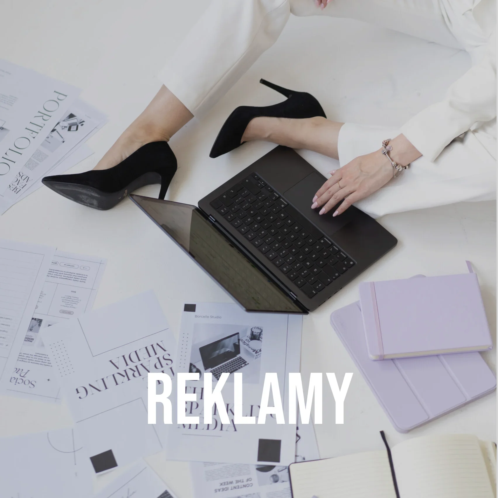 Dagmara Grzeszczuk z The Sparkling Media od marketingu dla architektów siedzi na ziemi w czarnych szpilkach. Obok leży laptop i kartki z logo firmy. Na środku biały napis "REKLAMY".